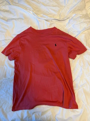 Röd t-shirt från Polo Ralph Lauren - Snygg röd t-shirt från Polo Ralph Lauren med klassisk rund hals och den ikoniska lilla loggan broderad på bröstet. Tillverkad i mjuk bomull som känns skön mot huden. Perfekt till jeans eller shorts för en clean och stilren look.