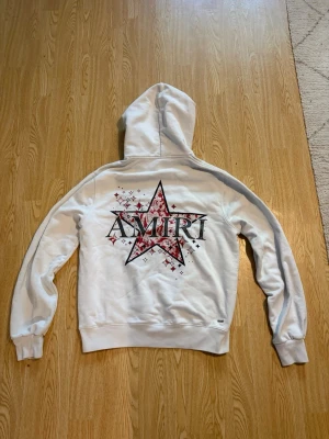 Vit hoodie med stjärntryck från Amiri - Säljer en vit hoodie från Amiri med stor stjärna och logga i svart och rött på ryggen samt mindre tryck på framsidan. Hoodien har huva, långa ärmar och en stor magficka. Materialet är mjukt och skönt, perfekt för en avslappnad streetwear-look.