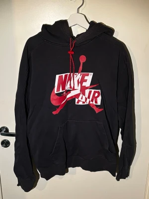 Svart Nike Air hoodie - Säljer en svart hoodie från Nike med stor röd och vit Nike Air-logga och Jumpman-tryck på bröstet i storlek M. Hoodien har huva med röda dragsnören och en klassisk magficka. Perfekt för dig som gillar streetwear och sportig stil.