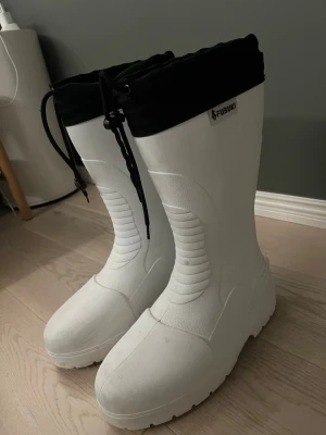 Vita höga stövlar från Fubuki - Säljer ett par vita höga stövlar från Fubuki med svart dragsko upptill. Stövlarna har en robust sula och är tillverkade i ett slitstarkt, vattentätt material. Perfekta för regniga dagar eller snöslask. Snygg och modern design som sticker ut.
