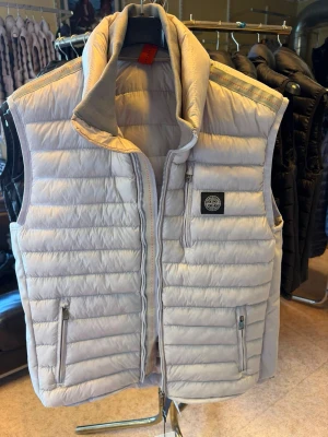 Ljusgrå dunväst från Stone Island - Snygg ljusgrå dunväst från Stone Island med quiltad design och hög krage. Västen har två dragkedjeförsedda fickor fram och en bröstficka med ikonisk patch. Tillverkad i lätt och värmande material, perfekt för lager-på-lager-stil.