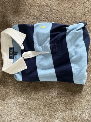 Randig rugbytröja från Polo Ralph Lauren - Säljer en klassisk långärmad rugbytröja från Polo Ralph Lauren med breda marinblå och ljusblå ränder, vit krage och knappslå. Tröjan har en gul broderad logga på bröstet och är gjord i mjuk bomull. Perfekt för en sportig och avslappnad stil.