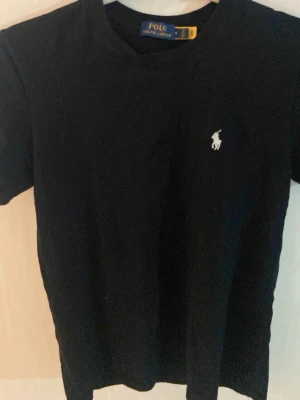 Svart t-shirt från Polo Ralph Lauren - Klassisk svart t-shirt från Polo Ralph Lauren med den ikoniska vita logobrodyr på bröstet. T-shirten har rund hals och korta ärmar, tillverkad i mjuk bomull för en skön känsla. Perfekt basplagg för en clean och stilren look.