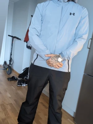 Ljusgrå vindjacka från Under Armour - Snygg ljusgrå vindjacka från Under Armour med dragkedja framtill, två fickor med dragkedja och justerbar huva. Jackan har en minimalistisk design med svart logga på bröstet och är tillverkad i ett lätt, vindtätt material. Använd ca tio ggr kvalite 8.5 av 10 så den ser väldigt fräsch ut inga tecken på användning ..då jag tyvärr e lite pedantisk och hålla saker på och runt mig :) st S passar M också 