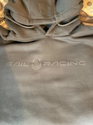 Grå hoodie från Sail Racing - Hoodie från Sail Racing med diskret logga på bröstet. Tillverkad i mjukt material och har en klassisk luva. Perfekt för en avslappnad och sportig stil.