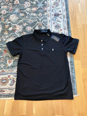 Svart pikétröja från Polo Ralph Lauren - Klassisk svart pikétröja från Polo Ralph Lauren med vit broderad logga på bröstet. Tröjan har korta ärmar, krage och tre knappar framtill. Perfekt till både jeans och shorts för en clean och stilren look.