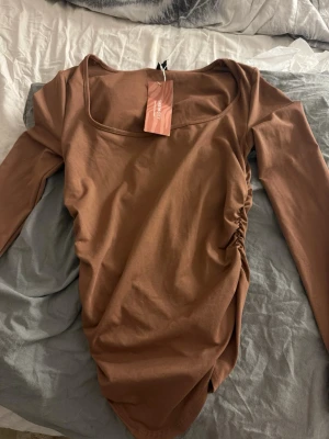 Brun långärmad topp från SHEIN - Snygg brun långärmad topp från SHEIN med fyrkantig ringning och rynkade detaljer på sidan. Tillverkad i mjukt stretchigt material som sitter bekvämt och har en modern, figurnära passform. Perfekt att styla med jeans eller kjol.