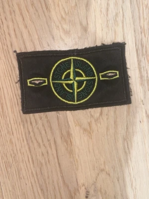 Stone Island tygmärke svart/gult - Säljer ett klassiskt Stone Island tygmärke i svart med broderad gul och grön kompasslogga. Märket har två knapphål på sidorna och är perfekt att sy eller fästa på jackor, tröjor eller väskor för en streetwear-look.