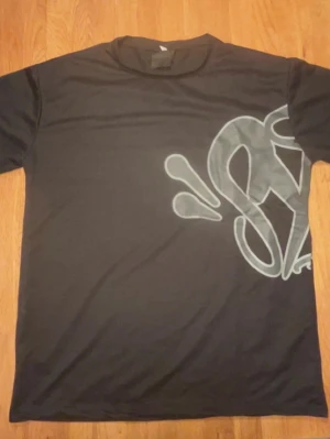 Syna World svart t-shirt - Snygg svart t-shirt med stor grå graffiti-inspirerad print som täcker både fram- och baksidan. T-shirten har rund hals och korta ärmar. Perfekt för dig som gillar streetwear och vill sticka ut med en unik design.
