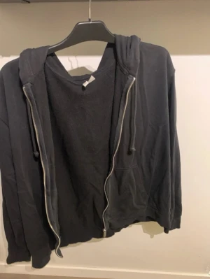 Svart zip hoodie med huva - En klassisk svart hoodie med dragkedja och huva. Hoodien har fickor framtill och justerbara snören i huvan. Tillverkad i mjukt bomullsliknande material och passar perfekt för en avslappnad stil.