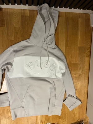 Ljusgrå/beige  hoodie från Hugo Boss - Snygg ljusgrå/beige hoodie från Hugo Boss med stor logga i relief på bröstet. Hoodien har en stor ficka framtill, dragsko i huvan och breda muddar vid ärmsluten. Tillverkad i mjukt bomullsmaterial för en skön och avslappnad look. 100% äkta pris✅ pris kan diskuteras😊