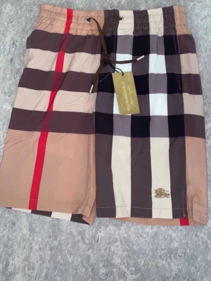 Burberry shorts - Säljer ett par Burberry shorts med det ikoniska mönstret i beige, svart, vitt och rött. Shortsen har snörning i midjan och en bakficka med Burberry-logga. Perfekta för sommaren och är riktigt schyssta 