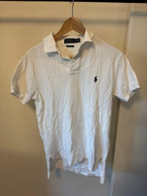 Vit pikétröja från Polo Ralph Lauren - Klassisk vit pikétröja från Polo Ralph Lauren med korta ärmar och den ikoniska mörkblå logotypen broderad på bröstet. Tröjan har krage och knappar vid halsen. Tillverkad i mjukt bomullsmaterial för en bekväm känsla.