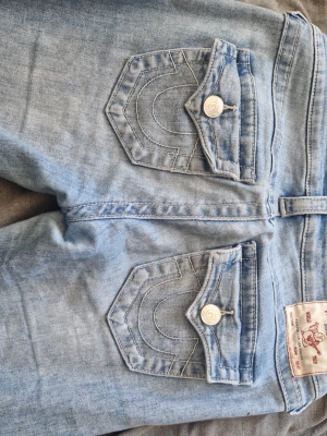 Ljusblå True Religion jeans, low rise - Tar så mycket du kan ge bytter också mot 26w i samma färg och modell Säljer ett par ljusblå jeans från True Religion med låg midja och utsvängda ben. Jeansen har ikoniska bakfickor med lock och vita kontrastsömmar samt snygga metallknappar. Perfekta för dig som gillar en avslappnad men trendig stil.