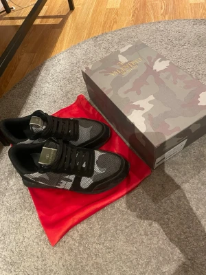 Valentino svart/gråa sneakers - Snygga sneakers från Valentino i svart och grått med kamouflagemönster. Skorna har en mix av mocka och meshmaterial, samt svarta snören och detaljer. Kommer med originalkartong och röd dustbag från Valentino Garavani.