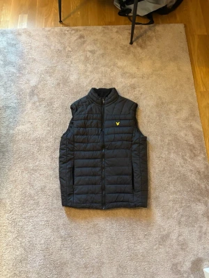 Svart dunväst med gul logga - Snygg svart dunväst med quiltad design och gul logga på bröstet. Perfekt för lager-på-lager och ger en clean look till din outfit.