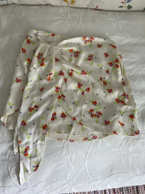 Vit omlottkjol med blommigt mönster - Söt vit omlottkjol med blommigt mönster i rött, grönt och lila. Kjolen har knytband i midjan och är tillverkad i ett lätt, flowy, silkes material som passar perfekt till varma dagar. Den har en feminin och somrig vibe med fina detaljer. Använd 1 gång.