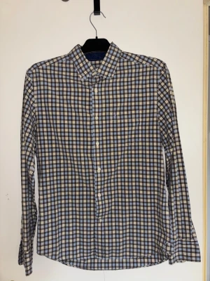 Peak Performance Skjorta - Snygg långärmad skjorta med rutigt mönster i blått, beige och vitt. Klassisk krage och knäppning framtill. Tillverkad i ett lätt bomullstyg som känns skönt mot huden. Perfekt för dig som gillar en avslappnad men ändå stilren look.