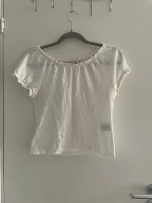 Vit kortärmad blus med volangkant - Söt vit blus med korta ärmar och volangkant vid halsringningen. Toppen har en lätt skrynklig struktur och är figurnära med en feminin touch. Perfekt att styla med jeans eller kjol för en fräsch look.