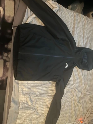 Svart vindjacka från The North Face - Snygg svart vindjacka från The North Face med huva och dragkedja framtill. Jackan har en minimalistisk design med logga på bröstet och ryggen. Perfekt för blåsiga dagar och enkel att matcha med olika outfits. Ny pris 1400 använd fåtal gånger 