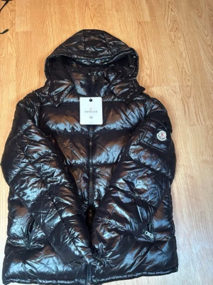 Svart dunjacka med huva från Moncler - Snygg svart dunjacka från Moncler med glansig finish och huva. Jackan har dragkedja framtill, två fickor med dragkedja och Moncler-logga på ärmen. Tillverkad i certifierat dun enligt DIST-standarden. Perfekt för kalla dagar.