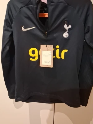 Tottenham Hotspur träningströja Nike - Mörkblå långärmad träningströja från Nike med Tottenham Hotspur-logga på bröstet och gul 'getir'-logga. Tröjan har halv dragkedja framtill och är tillverkad i ett funktionsmaterial som passar perfekt till träning. (XS junior)
