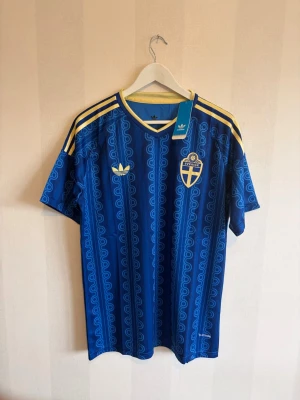 Blå Sverige fotbollströja Adidas - Säljer en blå fotbollströja från Adidas med Sveriges landslagsemblem på bröstet och gula detaljer på axlar och krage. Tröjan har ett unikt mönster i blå nyanser och texten 'Sverige' i nacken. Perfekt för match eller träning!