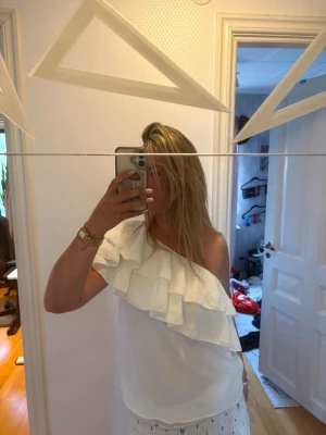 Vit oneshoulder topp med volang - Säljer en snygg vit oneshoulder topp med bred volangdetalj över axeln. Toppen är ärmlös på ena sidan och har en luftig passform, perfekt för sommarens alla tillfällen. Stilren och trendig design som passar till mycket.