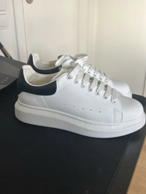 Alexander McQueen vita sneakers ( Aldrig använda ) - Stilrena vita sneakers från Alexander McQueen med svart hälparti och guldfärgad logga. Skorna har tjock sula, vita skosnören och diskreta ventilationshål på sidorna. Tillverkade i läder med klassisk låg modell och modern chunky design.