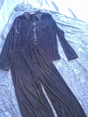 Plisserat svart loungewear-set - Setet innehåller skjorta och byxor. Materialet är glansigt och mjukt, perfekt för chill dagar hemma 🖤  Mycket bra skick och synes ej igenom, dubbla tyg på byxorna (sista bilden). Finns ett minihål som jag har råkat tappat rökelse på och ej synes när man har på sig det (näst sista bilden). 
