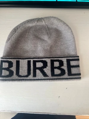 Grå mössa med svart Burberry-tryck - Säljer en stilren grå mössa från Burberry med bred uppvikt kant och stor svart logotyp runt hela mössan. Tillverkad i mjukt stickat material som håller dig varm och snygg under kalla dagar. Perfekt accessoar för dig som gillar klassisk design med en twist.