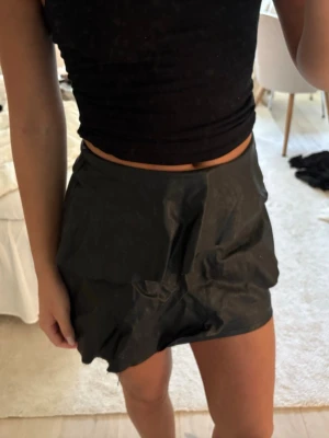 Svart kort kjol i skinnimitation - Säljer en svart kort kjol i skinnimitation med enkel design och hög midja. Kjolen har en lätt glansig yta och sitter snyggt på kroppen. Perfekt att matcha med en crop top eller stickad tröja. Storlek 146/152 men passar mig som är en xs/s