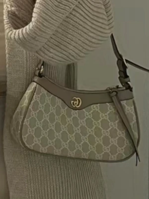 Beige axelväska med GG-mönster - Snygg beige axelväska med klassiskt GG-mönster och bruna detaljer. Väskan har guldfärgad logga framtill och justerbar axelrem. Perfekt storlek för det viktigaste och ger en trendig touch till din outfit.