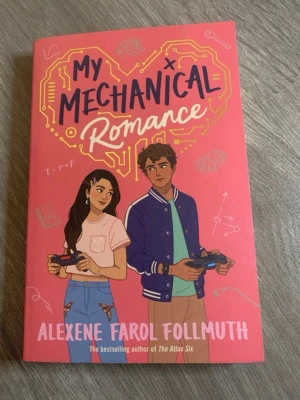 My Mechanical Romance bok - En ungdomsroman med rosa omslag och illustration av två tonåringar med spelkontroller. Titeln är tryckt i svart och vitt med ett hjärta i bakgrunden. Författare: Alexene Farol Follmuth. Perfekt för dig som gillar romantik och teknik.