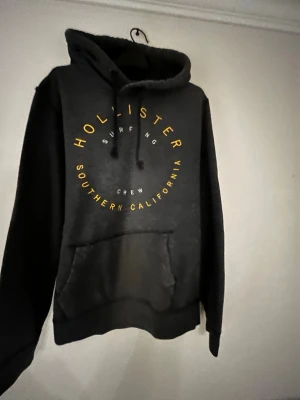 hoodie från Hollister - Mysig mörkgrå hoodie från Hollister med gul text på bröstet där det står 'Hollister Surfing Crew Southern California'. Hoodien har magficka, snörning i huvan och är långärmad. Perfekt för chill dagar och streetwear vibes.