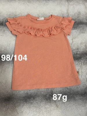 Aprikosfärgad topp med volang - Supersöt aprikosfärgad topp med korta ärmar och volangdetalj över bröstet. Perfekt för dig som gillar en gullig och enkel stil. Tillverkad i mjukt material som känns skönt mot huden.🌸 Fint använt skick. Färgen är typ orange/aprikos. Storlek 98/104