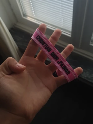 Rosa tygarmband med tofsar - Säljer ett rosa armband i tyg med texten 'Christian Dior J'Adior' i svart. Armbandet har dekorativa tofsar i ändarna och är justerbart för att passa olika handleder. Perfekt accessoar för att ge din outfit en färgklick.
