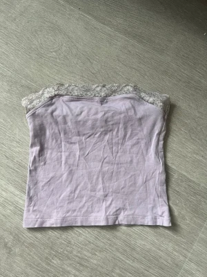 Ljuslila tubtopp med spetskant - Söt ljuslila tubtopp med dekorativ spetskant upptill. Toppen är axelbandslös och har en enkel, rak passform som känns både trendig och bekväm. Perfekt att styla till jeans eller kjol för en fräsch look.