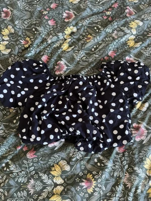 Svart prickig offshoulder topp - Säljer en svart offshoulder topp från Meet Me There med vita prickar och puffiga ärmar. Toppen har resår vid axlarna och en croppad passform som ger en trendig look! Aldrig använd!☺️☺️