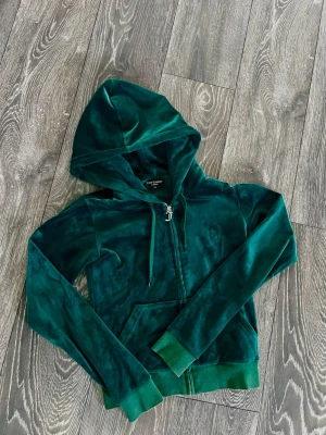 Grön velour hoodie med dragkedja - Mysig grön hoodie i mjuk velour med dragkedja framtill och broderad detalj på bröstet. Tröjan har huva med dragsko och ribbade muddar vid ärmslut och nederkant. Perfekt för en chill och avslappnad stil.