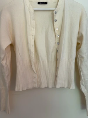 Vit ribbad kofta från Gina Tricot - Säljer en stilren vit kofta från Gina Tricot med ribbade detaljer vid ärmslut och nederkant. Koftan har långa ärmar och knäpps med silvriga knappar framtill. Perfekt att slänga över en topp för en enkel och clean look.
