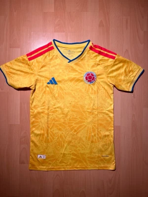 Colombia hemma matchtröja Adidas 2026 - Säljer en officiell Colombia matchtröja från Adidas i gult med röda och blå detaljer. Tröjan har korta ärmar, V-ringning med blå kant och röda ränder på axlarna. Tillverkad i lätt och ventilerande material, perfekt för fotboll eller träning.