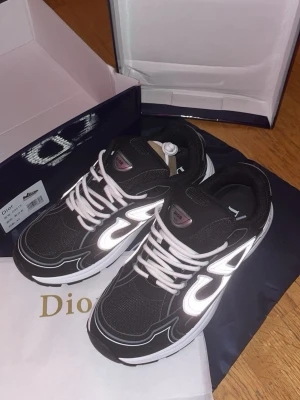 Svarta Dior sneakers med reflexdetaljer - Säljer ett par svarta Dior sneakers med vita och reflekterande detaljer. Skorna har vita snören, tjock vit sula och meshpartier för extra komfort. Snygg logga på sidan och tungspetsen. Perfekta för dig som vill sticka ut med exklusiv streetstyle.