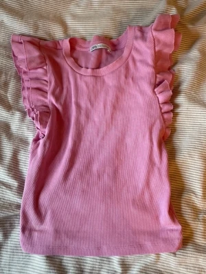 Topp från Zara - Supersöt rosa topp från Zara med ribbad struktur och volangdetaljer vid ärmarna. Modellen är ärmlös med rund halsringning och stretchigt material som sitter snyggt. Perfekt för dig som vill ha en feminin och trendig look.