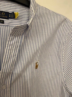 Randig skjorta från Polo Ralph Lauren - Klassisk skjorta från Polo Ralph Lauren i vitt och ljusblått med smala ränder. Skjortan har lång ärm, knappar framtill och den ikoniska broderade loggan på bröstet. Tillverkad i bomull och har en normal passform. Perfekt för en clean och stilren look.