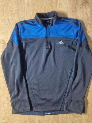 Mörkblå Adidas träningströja med dragkedja - Sportig långärmad träningströja från Adidas i mörkblått med blå axelpartier och klassiska ränder. Tröjan har halv dragkedja framtill och Adidas-logga på bröstet. Perfekt för träning eller uppvärmning. Tillverkad i funktionsmaterial.