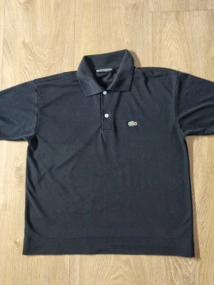 Svart pikétröja från Lacoste - Klassisk svart pikétröja från Lacoste med korta ärmar, krage och två knappar framtill. Ikoniska krokodilloggan på bröstet. Tillverkad i mjuk bomull som känns skön mot huden. OBS!! Jag har kortat ner den så skriv om ni vill ha måtten.