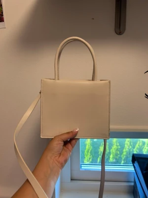 Beige fyrkantig handväska - Snygg beige handväska i fyrkantig modell med både handtag och axelrem. Väskan är gjord i slätt konstläder och har ett minimalistiskt uttryck. Perfekt för dig som gillar stilrena accessoarer och vill ha en väska som sticker ut lite extra.