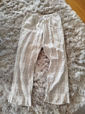 Rutiga pyjamasbyxor i bomull - Mysiga pyjamasbyxor i ljus bomull med rutigt mönster i vitt, grått och ljusrosa. Byxorna har resår i midjan och snörning för extra komfort. Perfekta för slappa dagar hemma eller en skön natts sömn.