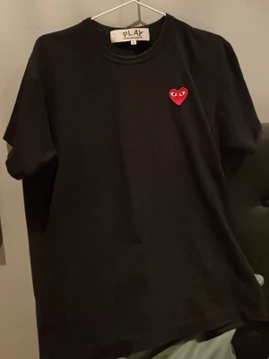 Svart Comme des Garçons PLAY t-shirt - Svart t-shirt från Comme des Garçons PLAY med det ikoniska röda hjärtat broderat på bröstet. Klassisk rund hals och korta ärmar. Tillverkad i mjuk bomull för en skön och avslappnad passform. Använd cirka 3 gånger men inga tecken på användning. Den är i storlek large och är liten i storleken. Bara höra av er om ni är intresserade:).